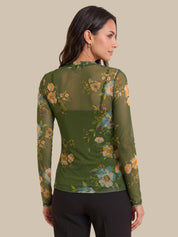 A woman shown from the back wearing a sheer olive green long-sleeve top with a floral print in yellow, blue, and white, paired with black pants, against a solid beige background.