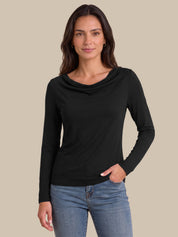 A woman modeling a black long-sleeve cowl neck top with blue jeans, set against a solid beige background.