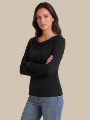 A woman modeling a black long-sleeve cowl neck top with blue jeans, standing against a solid beige background.