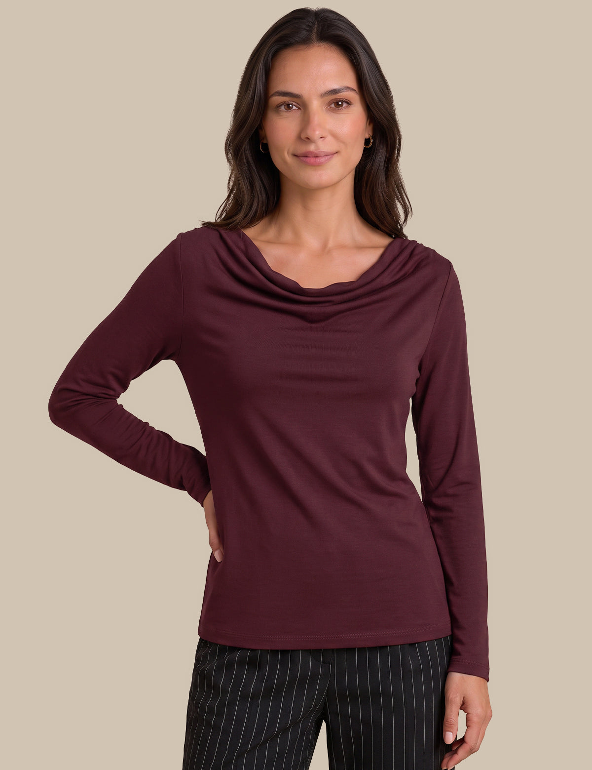 A woman modeling a long-sleeve cowl neck top in burgundy, paired with black pinstriped trousers, against a solid beige background.