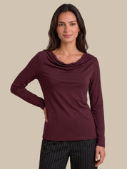 A woman modeling a long-sleeve cowl neck top in burgundy, paired with black pinstriped trousers, against a solid beige background.