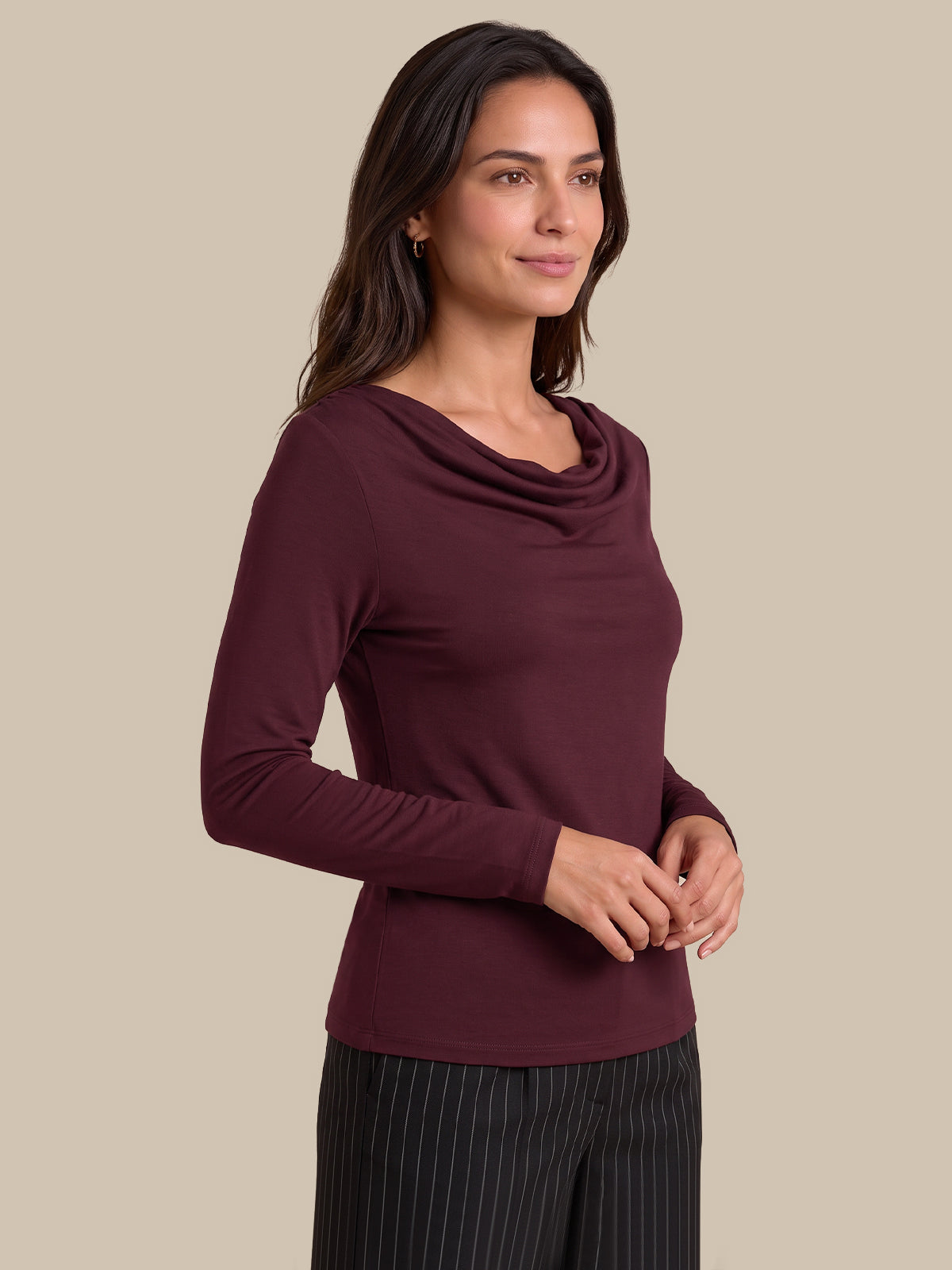 A woman modeling a long-sleeve cowl neck top in deep burgundy, paired with dark pinstriped trousers, against a solid beige background.