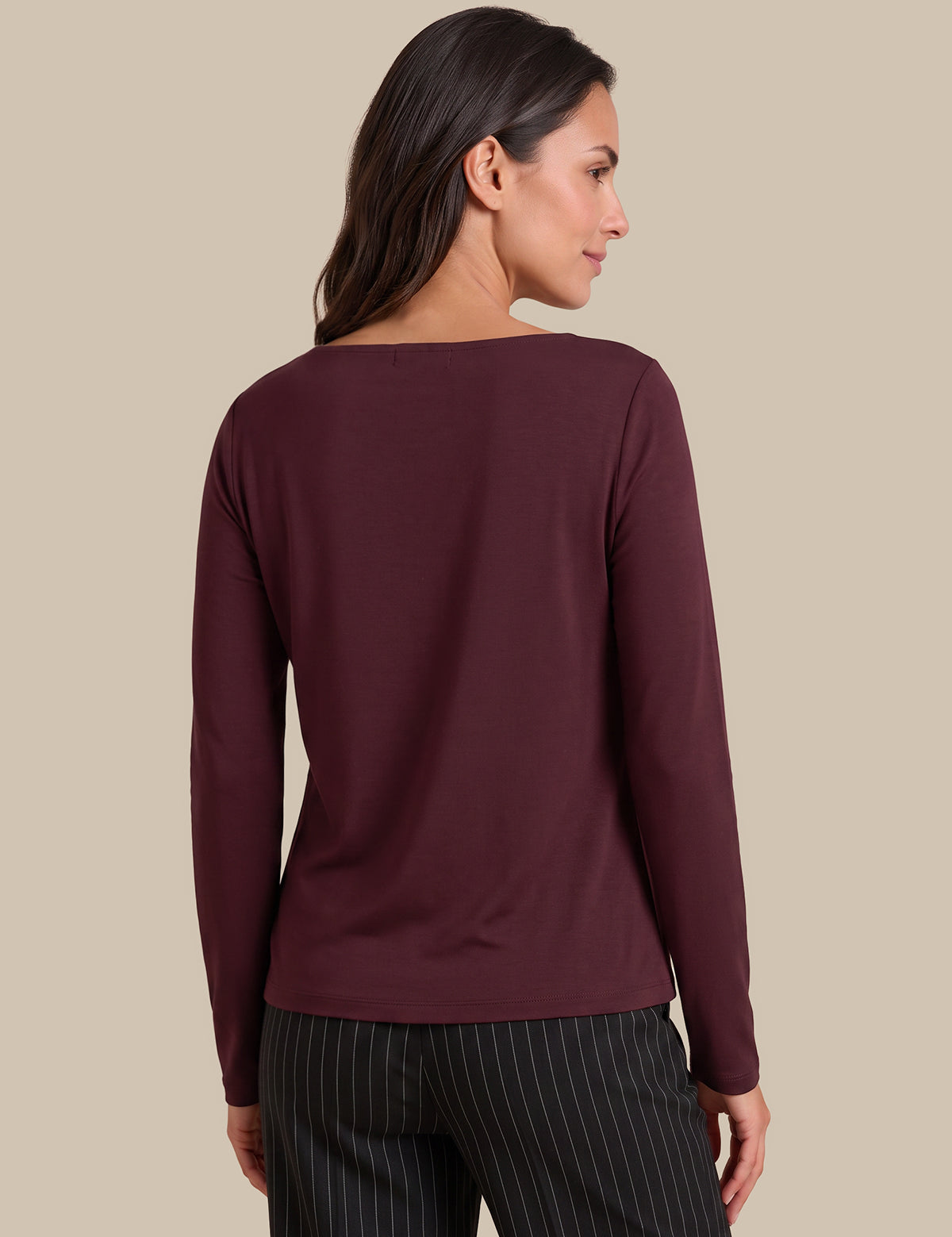 A woman shown from the back wearing a long-sleeve boat-neck top in deep burgundy, paired with dark pinstriped trousers, against a solid beige studio background.