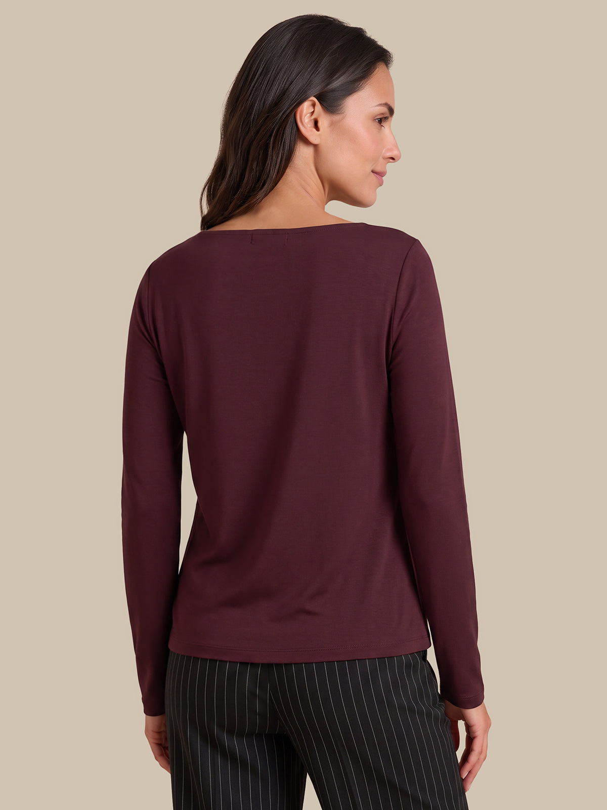 A woman shown from the back wearing a long-sleeve boat-neck top in deep burgundy, paired with dark pinstriped trousers, against a solid beige studio background.