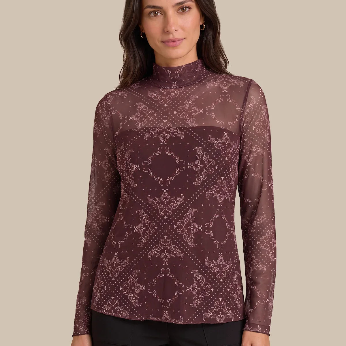 A woman modeling a long-sleeve mock neck top with a sheer mesh fabric and an intricate geometric damask pattern in dark burgundy and dusty rose, paired with black pants against a solid beige background.