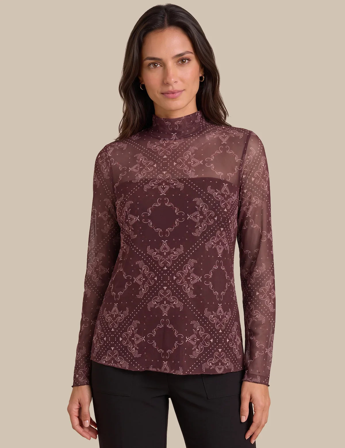 A woman modeling a long-sleeve mock neck top with a sheer mesh fabric and an intricate geometric damask pattern in dark burgundy and dusty rose, paired with black pants against a solid beige background.