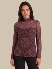 A woman modeling a long-sleeve mock neck top with a sheer mesh fabric and an intricate geometric damask pattern in dark burgundy and dusty rose, paired with black pants against a solid beige background.