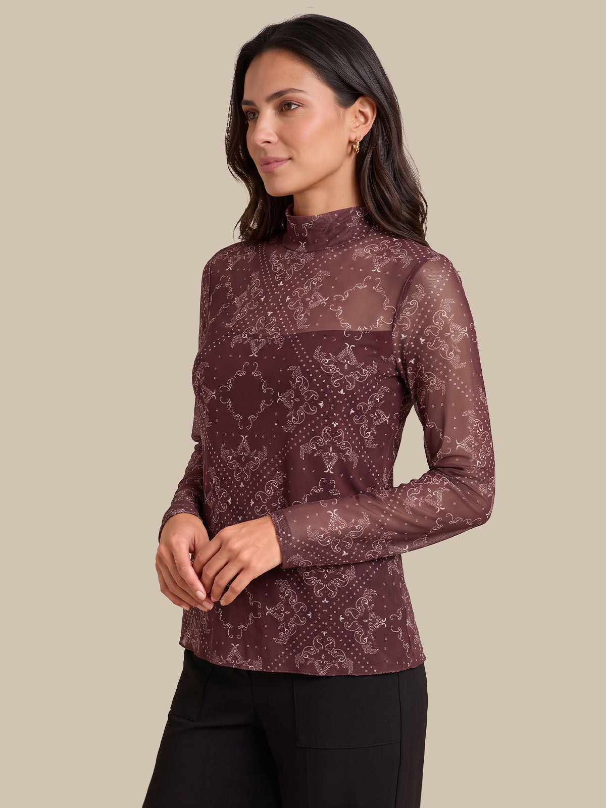 A woman modeling a long-sleeve mock neck top in a sheer maroon-brown color with a tonal floral lace pattern, paired with black pants, against a solid beige background.