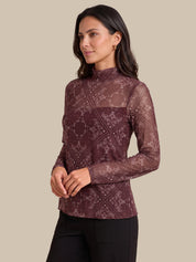 A woman modeling a long-sleeve mock neck top in a sheer maroon-brown color with a tonal floral lace pattern, paired with black pants, against a solid beige background.