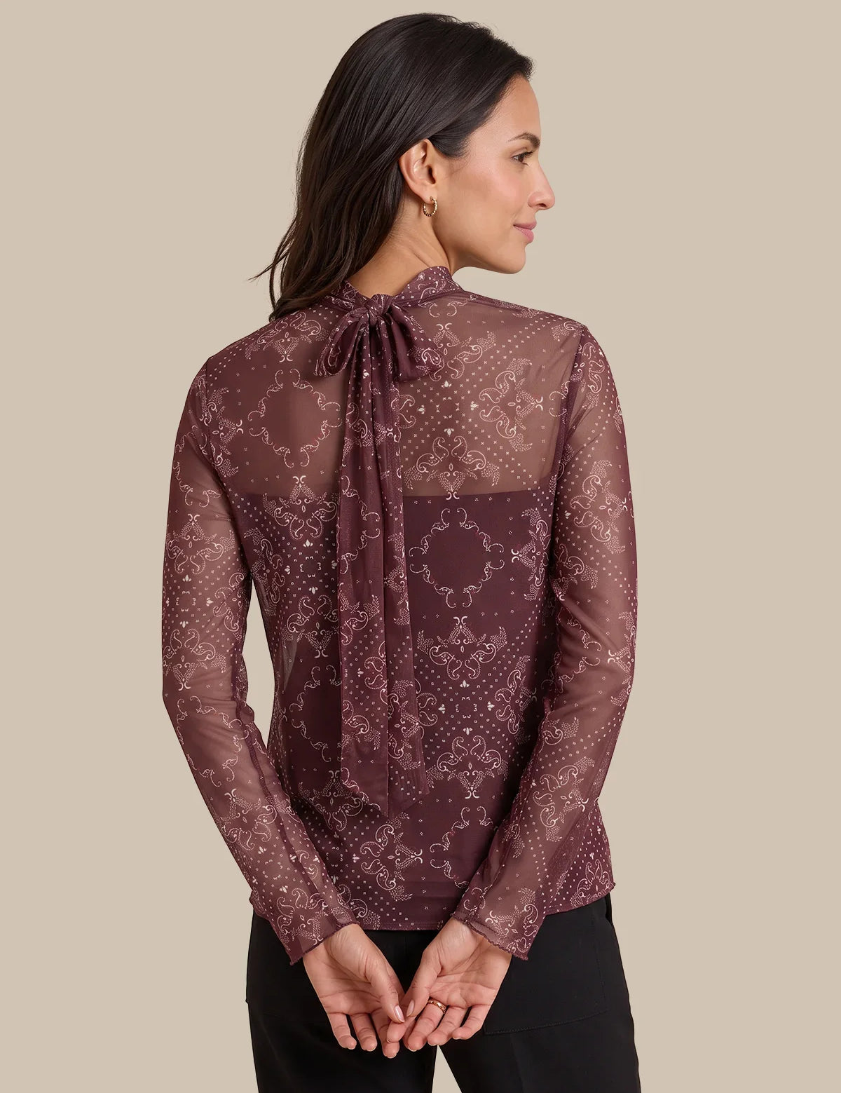 A woman shown from the back wearing a sheer long-sleeve blouse with an allover snakeskin pattern in dark brown and burgundy, featuring a tie-neck detail, paired with black pants against a solid beige background.