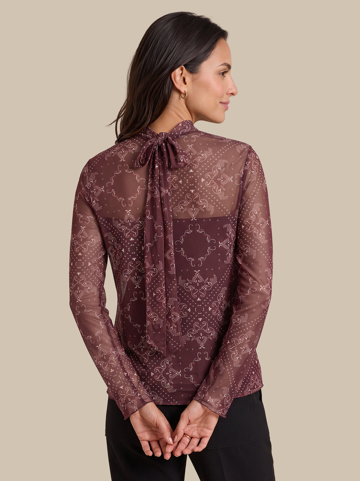 A woman shown from the back wearing a sheer long-sleeve blouse with an allover snakeskin pattern in dark brown and burgundy, featuring a tie-neck detail, paired with black pants against a solid beige background.