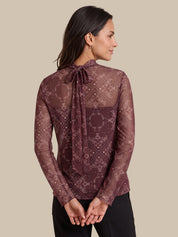 A woman shown from the back wearing a sheer long-sleeve blouse with an allover snakeskin pattern in dark brown and burgundy, featuring a tie-neck detail, paired with black pants against a solid beige background.