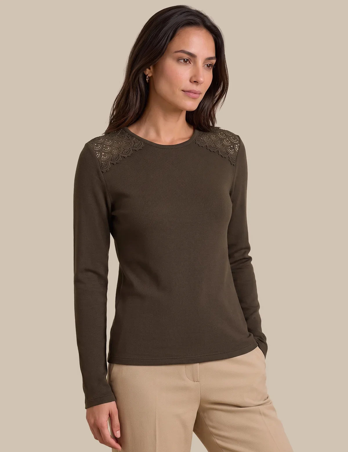 A woman modeling a dark brown long-sleeve knit top with lace shoulder detailing, paired with beige pants, against a solid beige background.