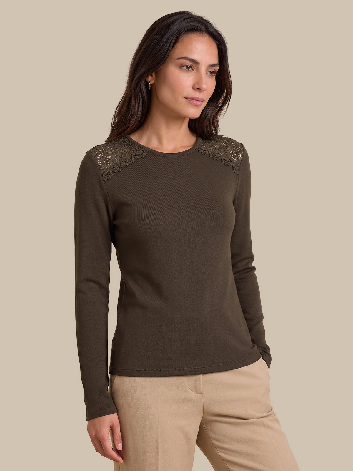 A woman modeling a dark brown long-sleeve knit top with lace shoulder detailing, paired with beige pants, against a solid beige background.
