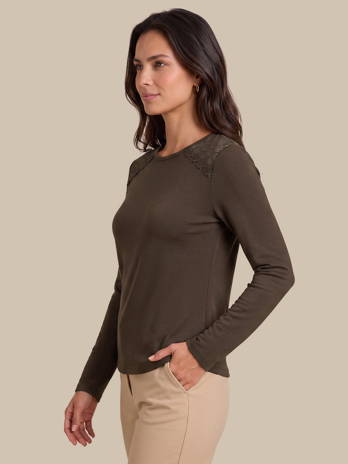 A woman wearing a dark brown long-sleeve knit top with lace detailing on the shoulders, paired with beige pants, photographed against a solid beige background.
