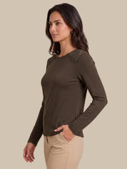 A woman wearing a dark brown long-sleeve knit top with lace detailing on the shoulders, paired with beige pants, photographed against a solid beige background.