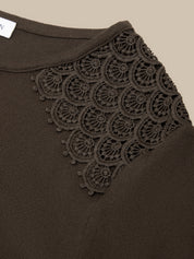 Close-up of a dark brown knit top featuring an intricate circular lace applique panel across the upper chest, set against a solid beige background.