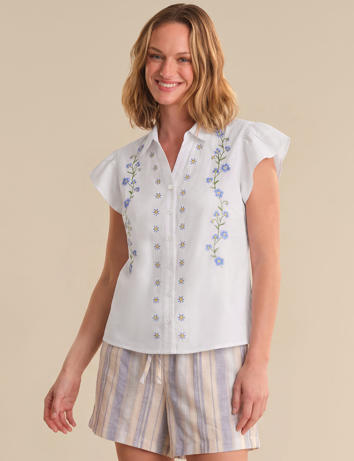 Woman wearing a white blouse with floral embroidery and striped shorts on a beige background