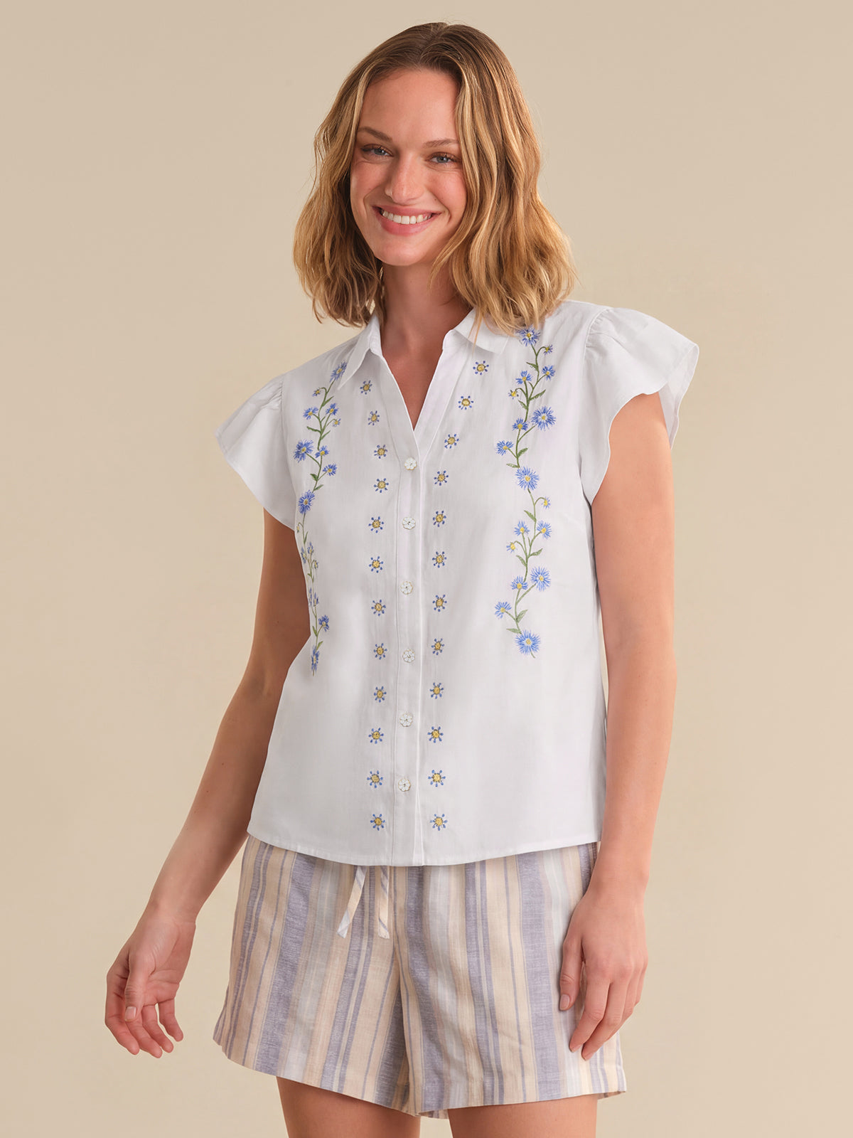 Woman wearing a white blouse with floral embroidery and striped shorts on a beige background