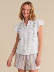 Woman wearing a white blouse with floral embroidery and striped shorts on a beige background