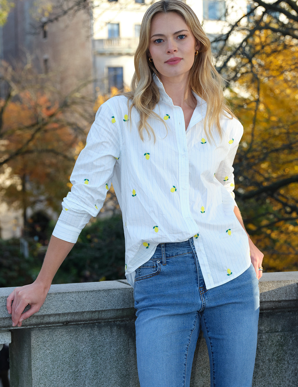 Woman wearing a white shirt with lemon patterns and blue jeans, standing outdoors with trees in the background.