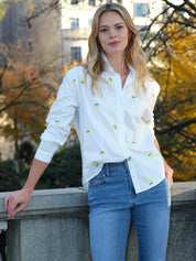 Woman wearing a white shirt with lemon patterns and blue jeans, standing outdoors with trees in the background.