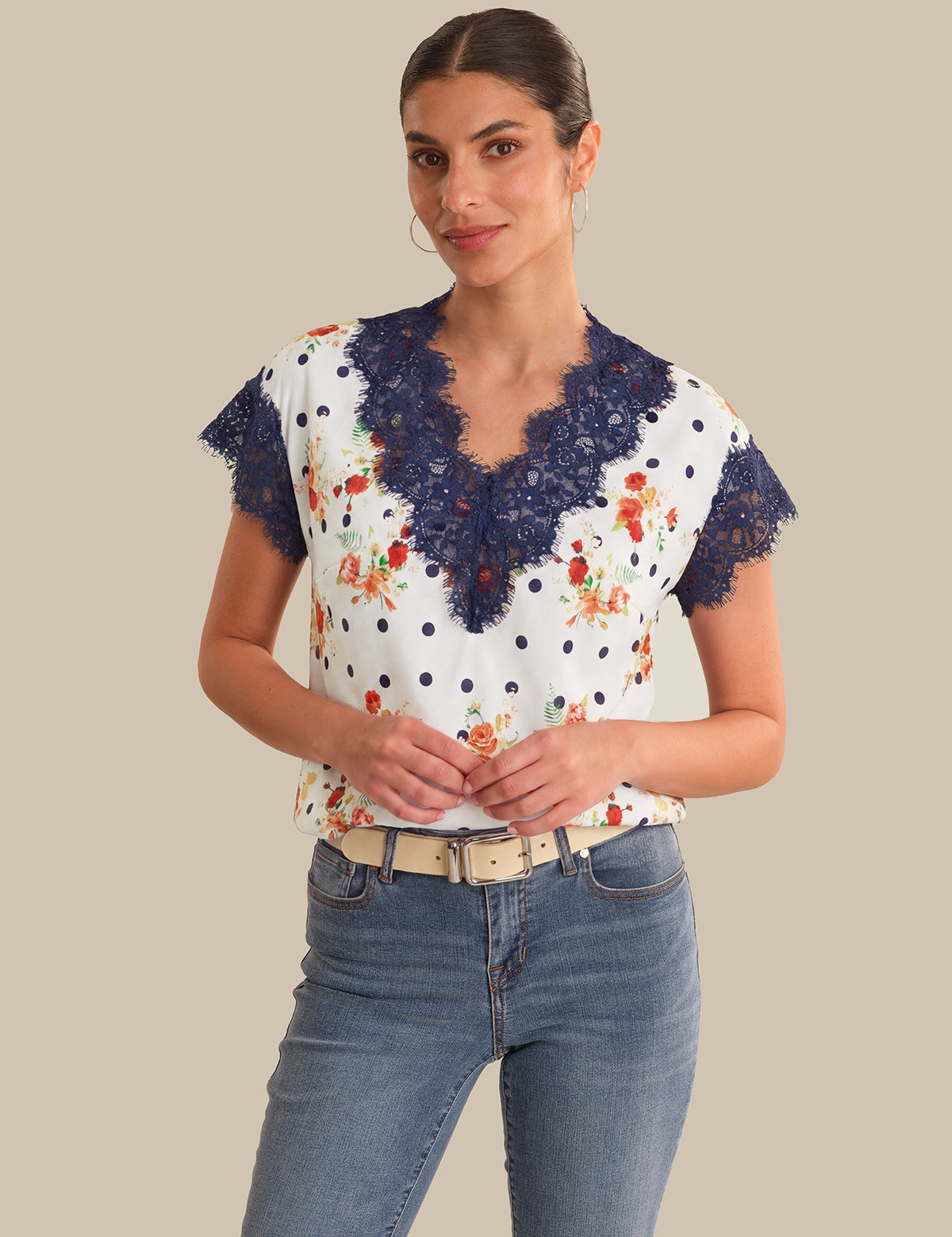 Woman wearing a white blouse with blue lace details and floral patterns, paired with blue jeans on a beige background.