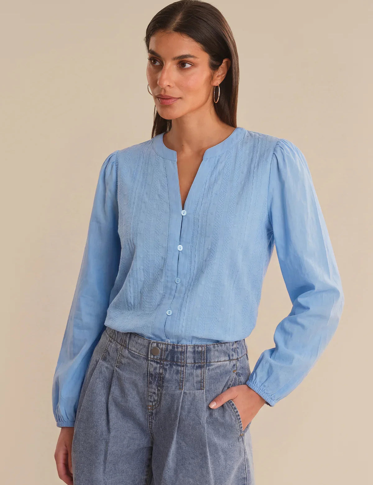 Woman wearing a light blue blouse with long sleeves and high collar, standing against a beige background.