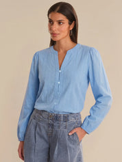 Woman wearing a light blue blouse with long sleeves and high collar, standing against a beige background.