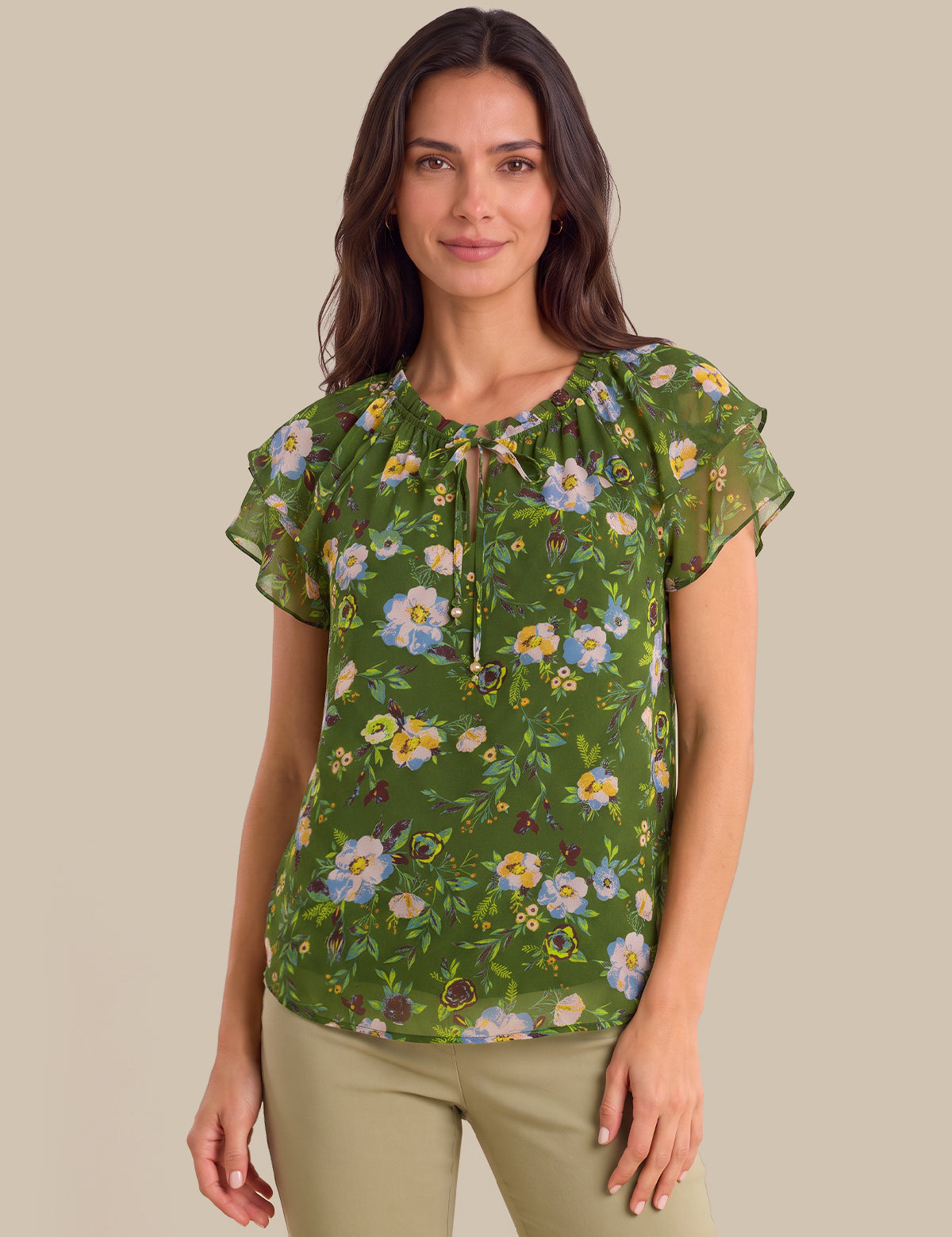 A woman modeling a green short-sleeve blouse with a floral print, featuring ruffled sleeves and a keyhole neckline with tie details, paired with khaki pants against a solid beige background.