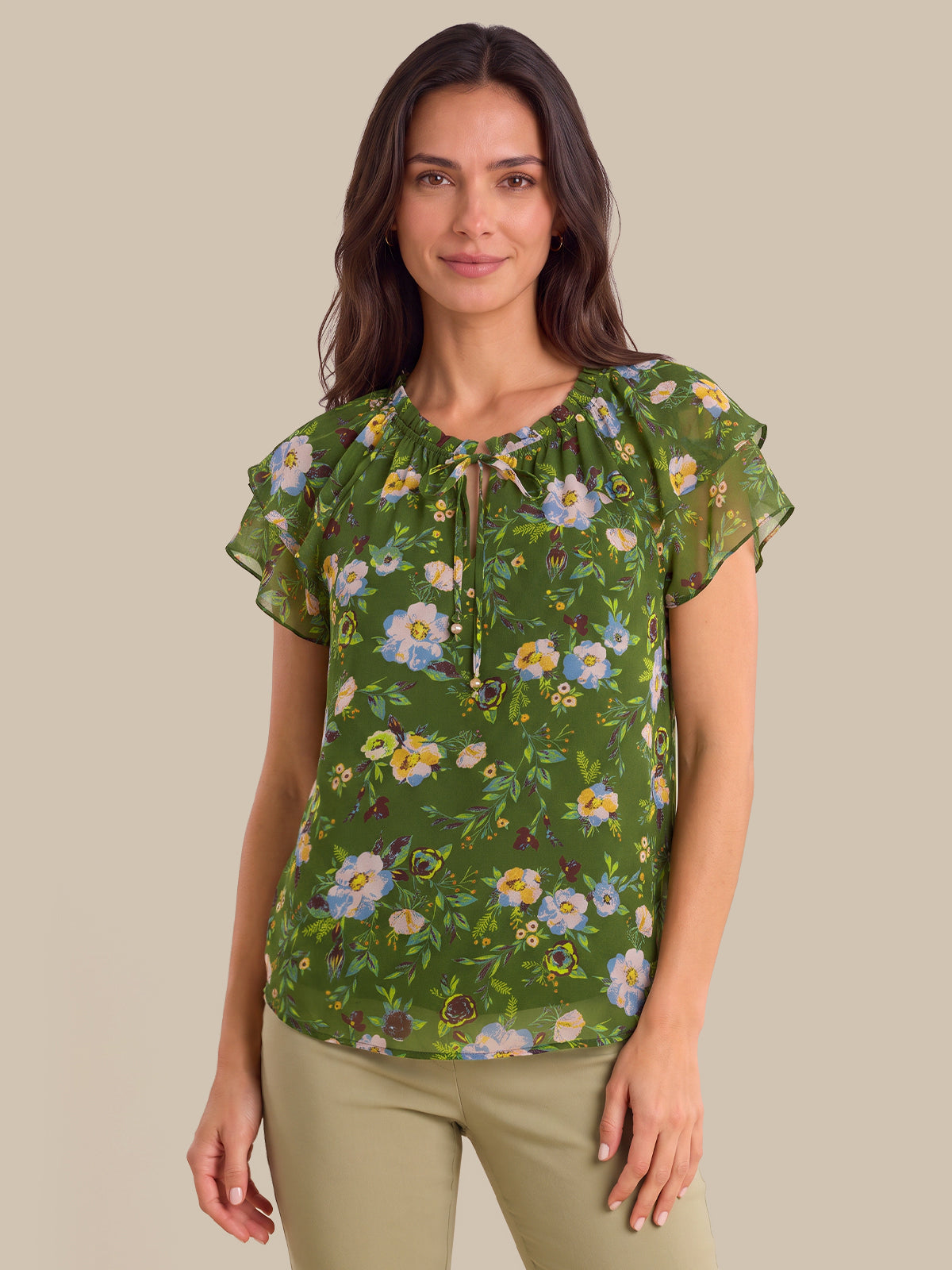 A woman modeling a green short-sleeve blouse with a floral print, featuring ruffled sleeves and a keyhole neckline with tie details, paired with khaki pants against a solid beige background.