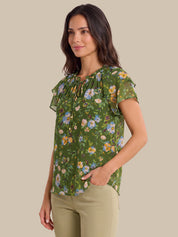 A woman wearing a green short-sleeve blouse with a colorful floral print and ruffled neckline, paired with beige pants, against a solid beige background.