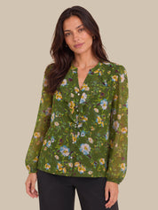 A woman wearing a green long-sleeve blouse with a yellow, blue, and white floral print, paired with black pants, against a solid beige background.