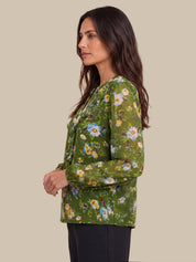A woman shown in side profile wearing a green long-sleeve blouse with a colorful floral print, paired with black pants, against a solid beige background.