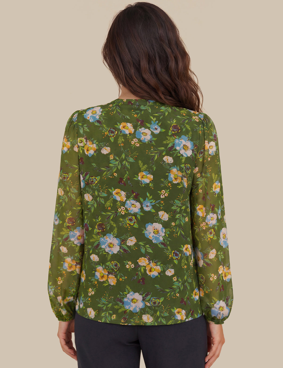 A woman viewed from the back wearing a green long-sleeve blouse with a colorful floral print and dark pants, set against a solid beige background.