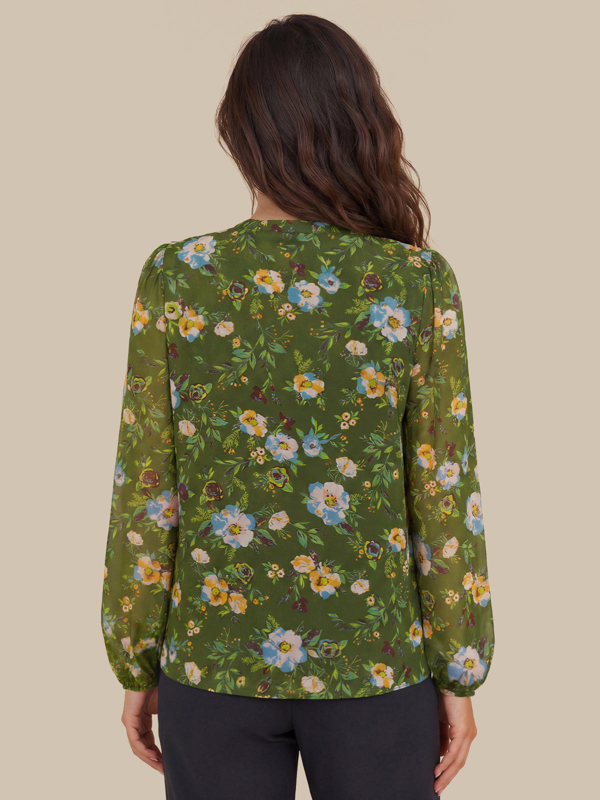 A woman viewed from the back wearing a green long-sleeve blouse with a colorful floral print and dark pants, set against a solid beige background.
