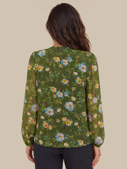 A woman viewed from the back wearing a green long-sleeve blouse with a colorful floral print and dark pants, set against a solid beige background.