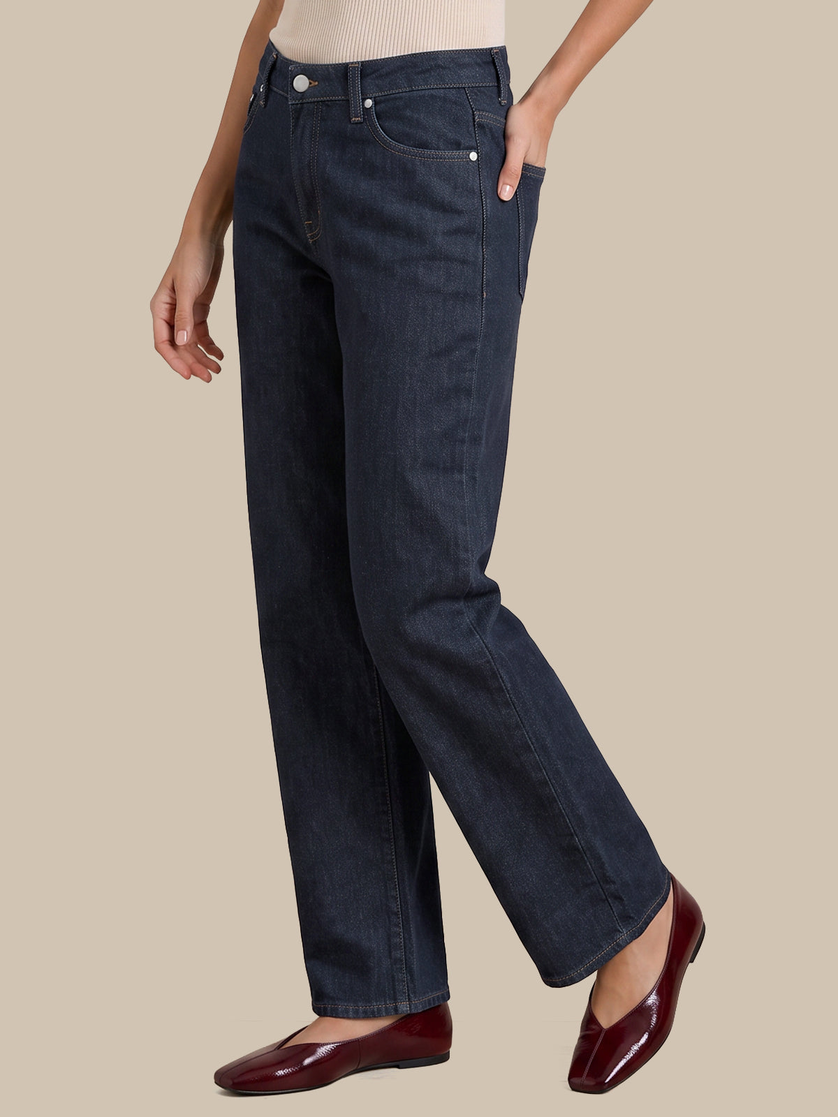 A person wearing dark blue straight-leg denim jeans, a cream ribbed top, and burgundy patent leather flats, standing against a solid beige background.