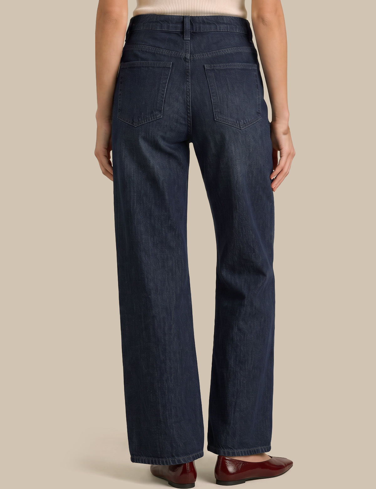 Rear view of a model wearing dark blue wide-leg denim jeans, paired with a light beige top and dark red flats, against a solid beige background.