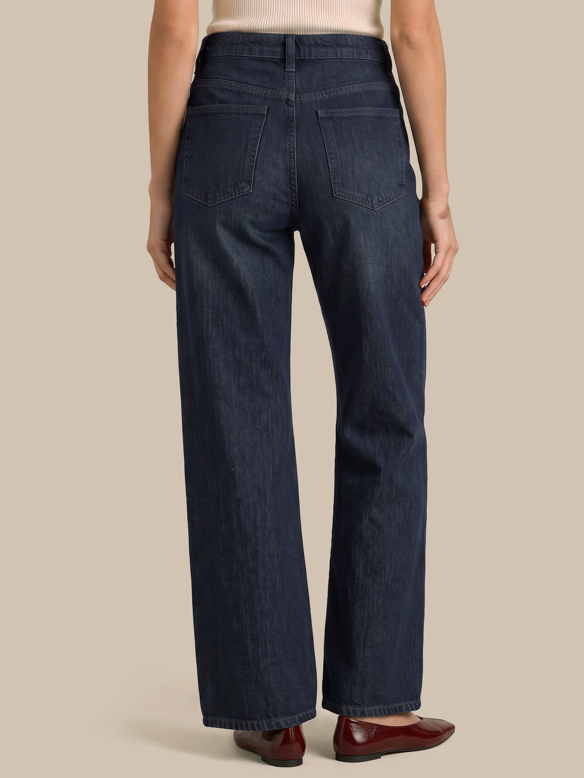Rear view of a model wearing dark blue wide-leg denim jeans, paired with a light beige top and dark red flats, against a solid beige background.