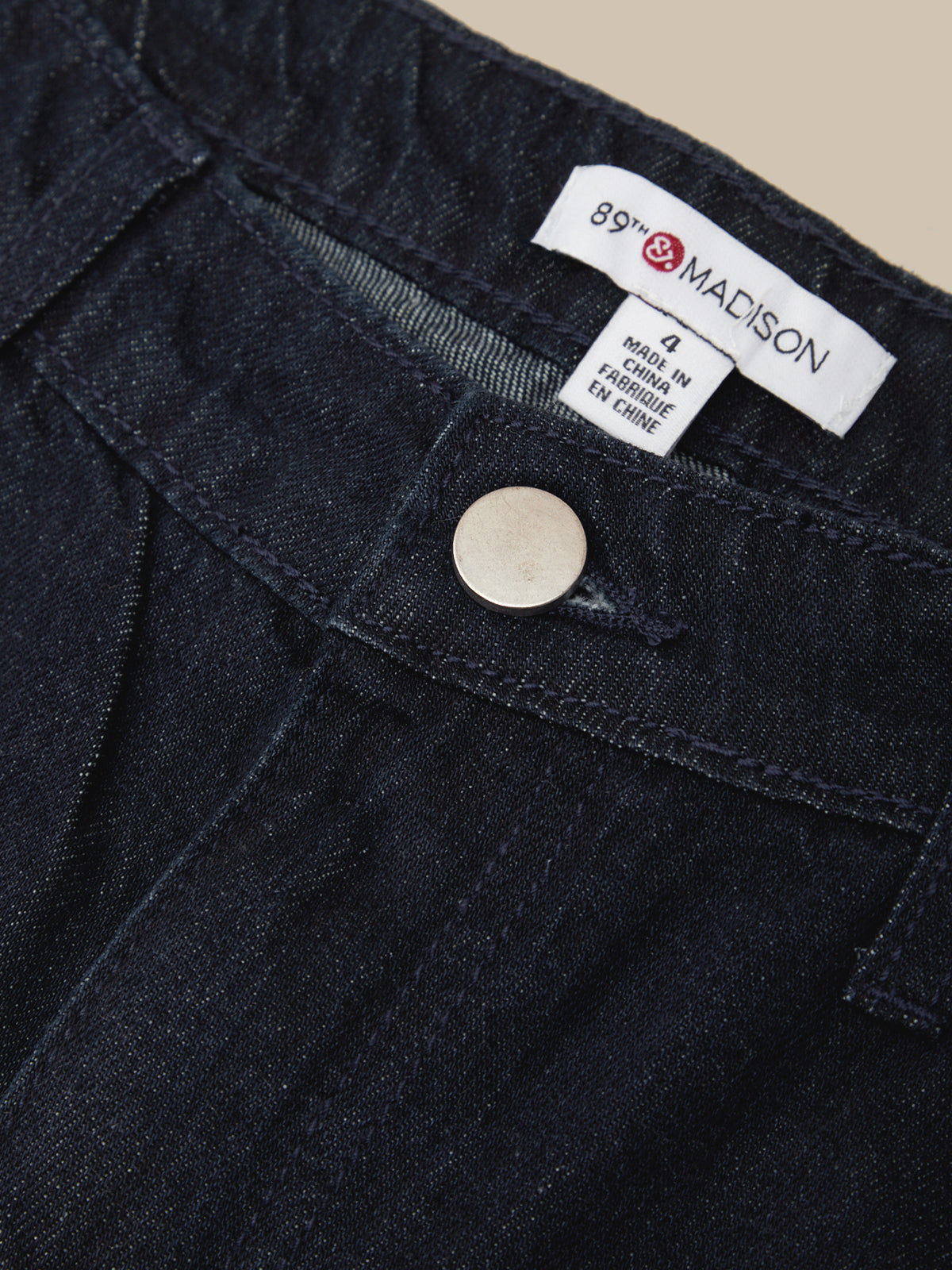 Close-up of a dark indigo denim garment, likely jeans or trousers, showing the waistband, button, and interior labels including the visible brand 89th Madison.
