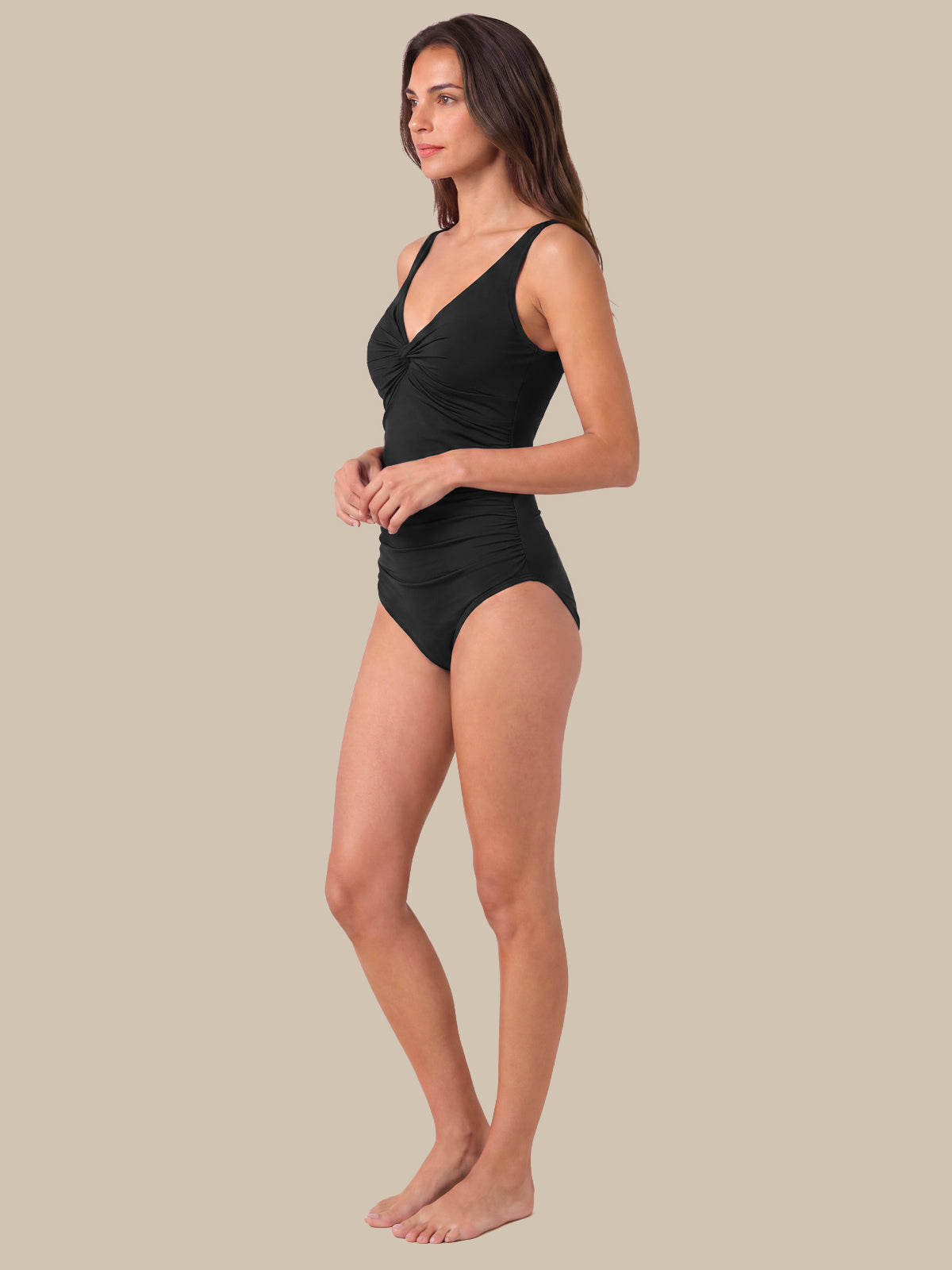 A woman modeling a black one-piece swimsuit in a side profile view against a solid beige background.
