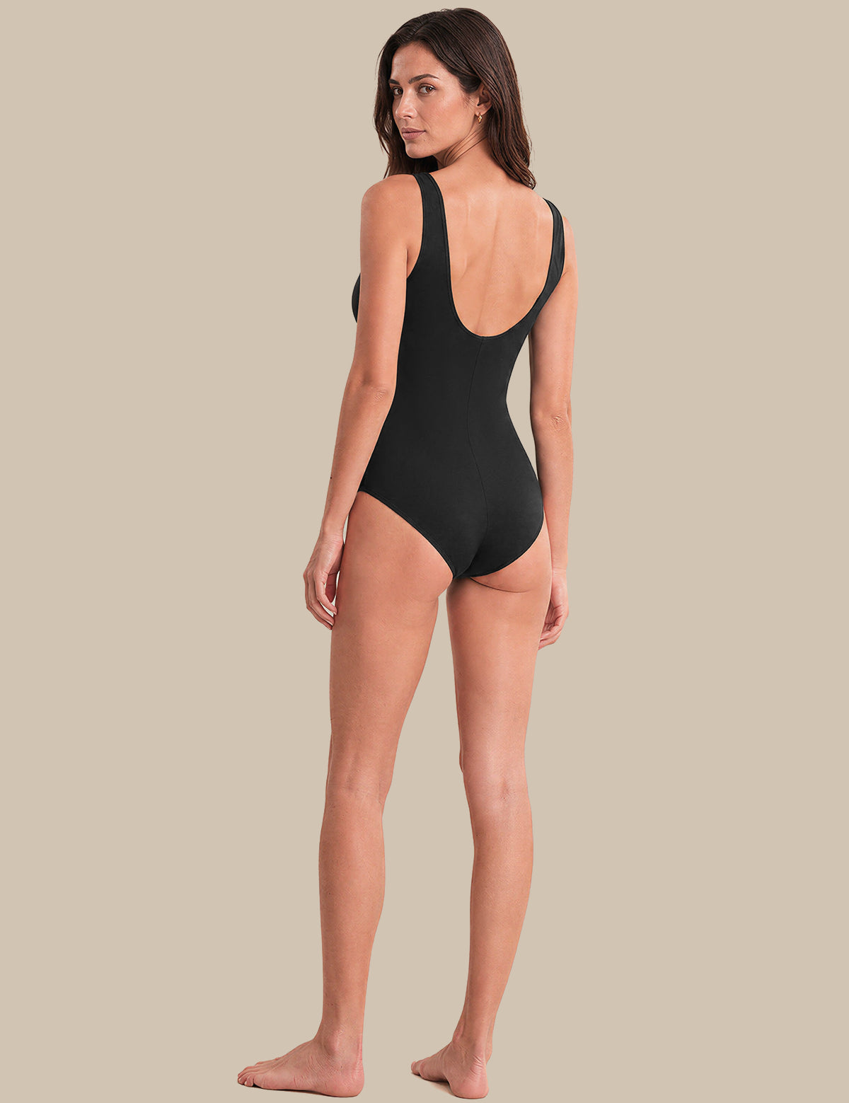 A woman modeling a black one-piece swimsuit from the back, showing a deep open back and shoulder straps, against a solid beige background.