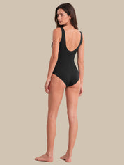 A woman modeling a black one-piece swimsuit from the back, showing a deep open back and shoulder straps, against a solid beige background.
