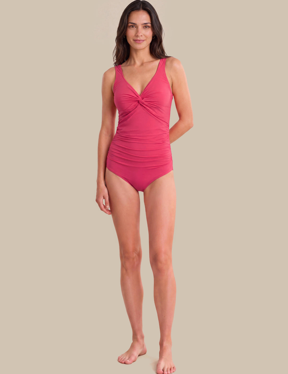 A woman modeling a bright pink one-piece swimsuit with a twisted front detail, standing against a solid beige background.