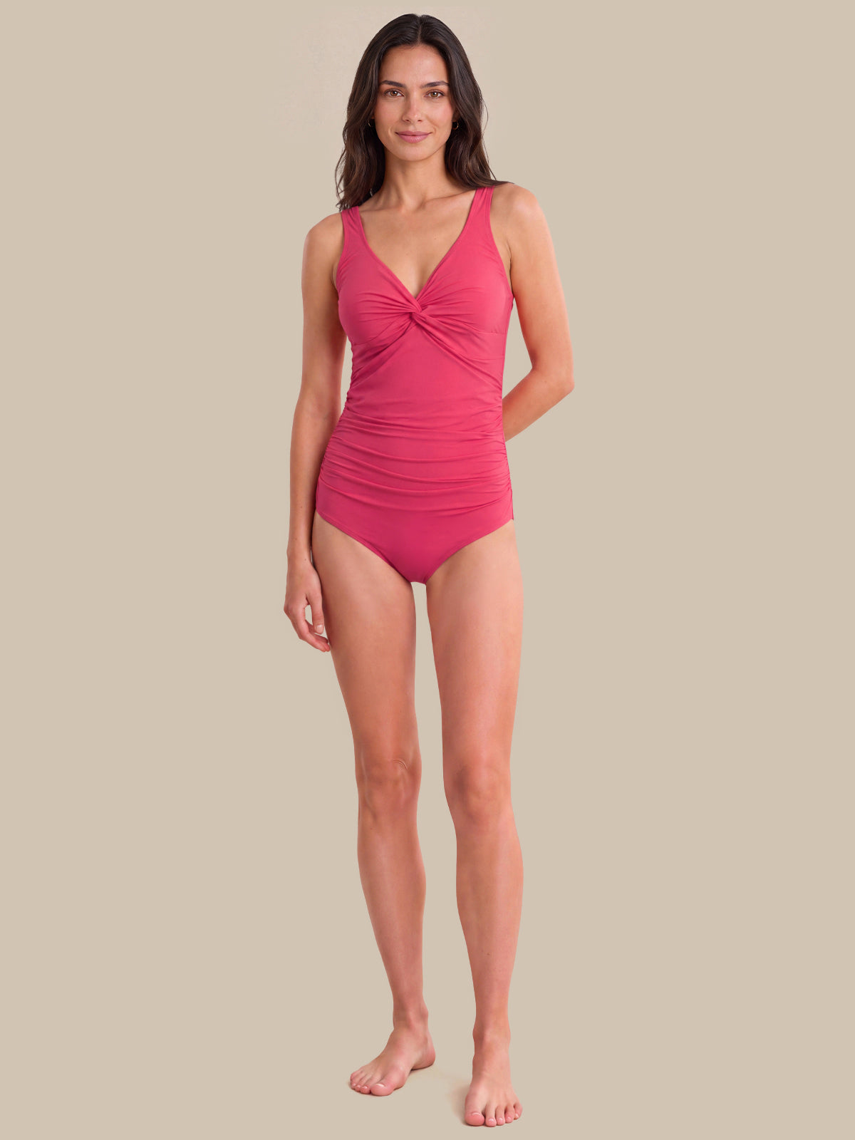A woman modeling a bright pink one-piece swimsuit with a twisted front detail, standing against a solid beige background.