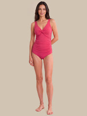 A woman modeling a bright pink one-piece swimsuit with a twisted front detail, standing against a solid beige background.