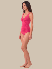 A woman modeling a bright pink one-piece swimsuit with a ruched front and V-neckline, shown in profile against a solid beige background.