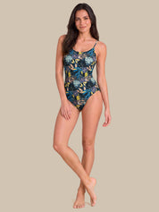 A woman modeling a one-piece swimsuit with a tropical floral print in shades of blue, green, and yellow on a dark background, set against a solid beige studio backdrop.