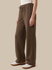 A person wearing loose, wide-leg brown drawstring pants with white sneakers, shown from the waist down against a plain beige studio background.